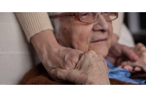 Ältere Frau sitz in einem Sessel und hält zufrieden die Händereiner Jüngeren, die sie an den Schultern berühren.