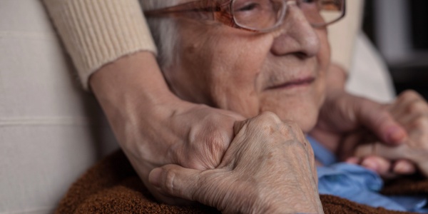 Ältere Frau sitz in einem Sessel und hält zufrieden die Hände einer Jüngeren, die sie an den Schultern berühren.